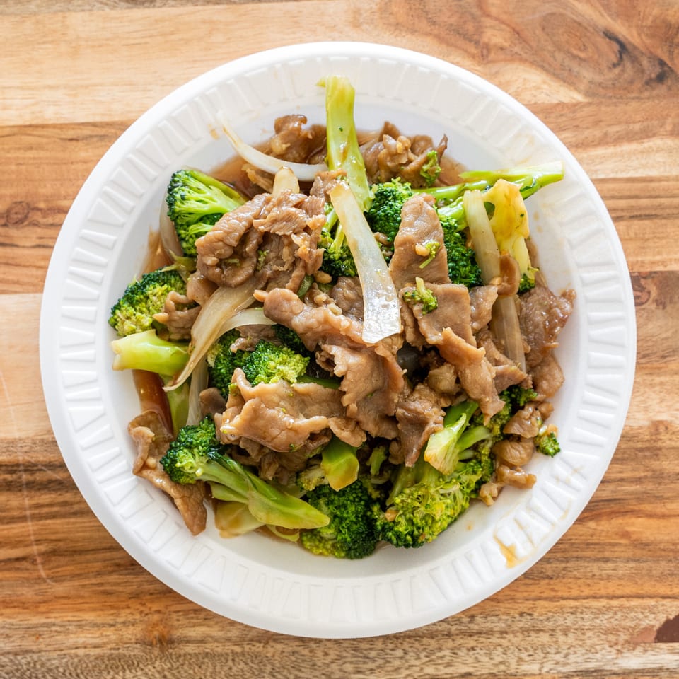 Stir-fried beef with broccoli, onions, and celery served in a white disposable bowl on a wooden table.