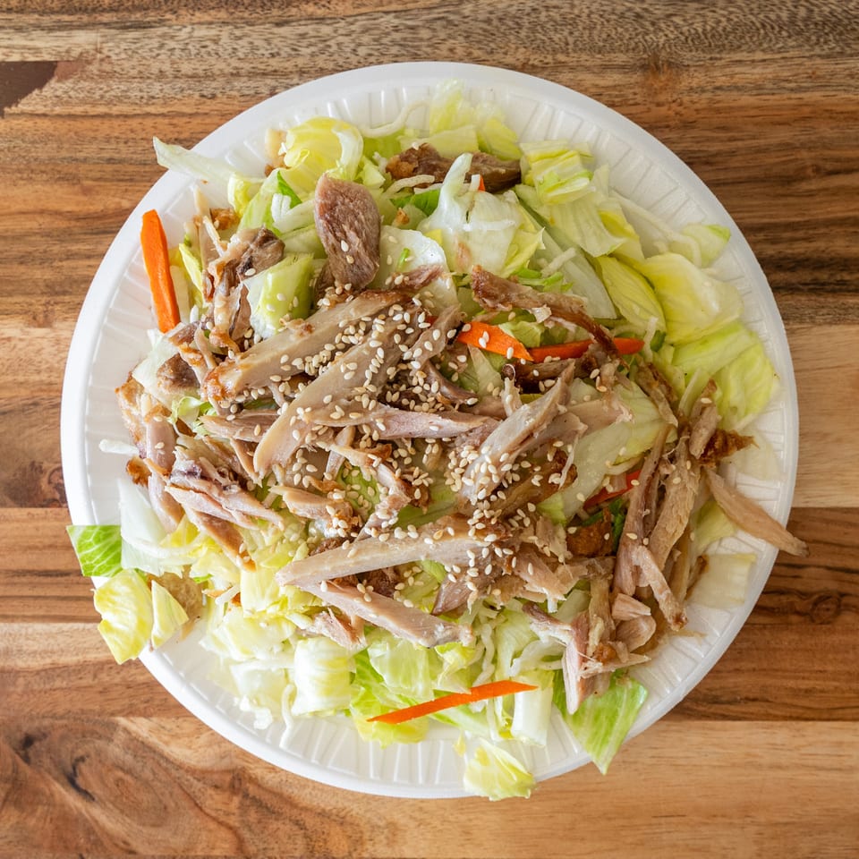 Lettuce salad topped with sliced roasted meat, carrots, and sesame seeds in a disposable bowl on a wooden table.