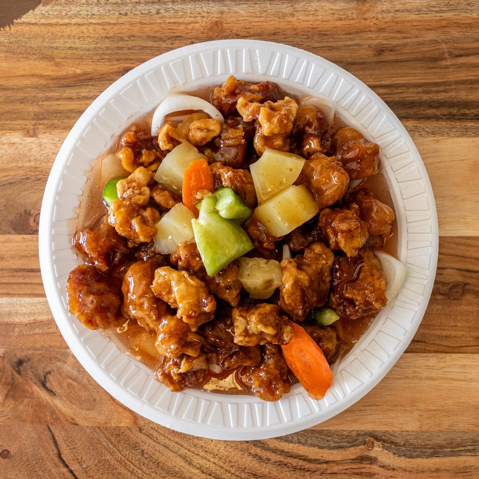 Sweet-and-sour battered chicken with pineapple, bell peppers, onions, and carrots in a white takeout plate on a wooden table.