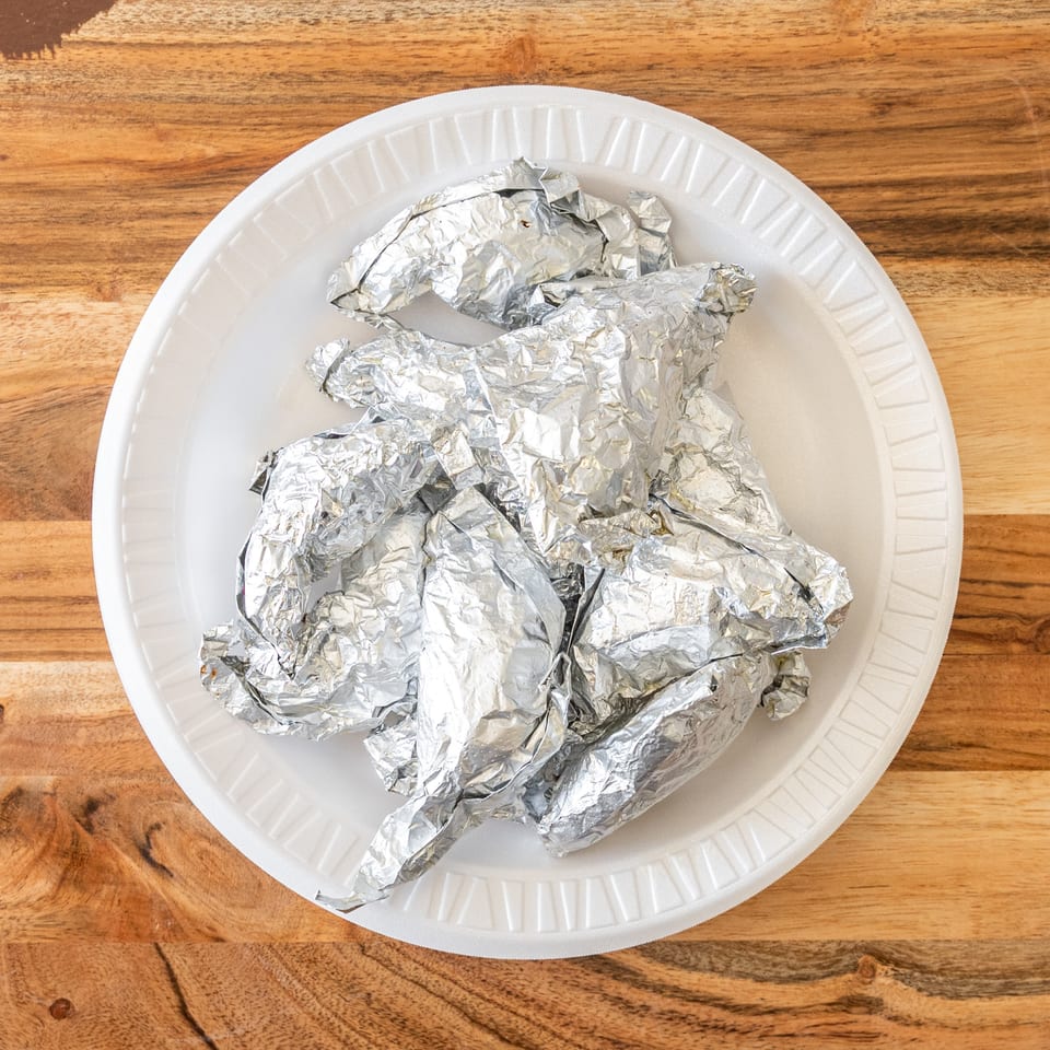 Several foil-wrapped food items arranged on a white disposable plate atop a wooden table.